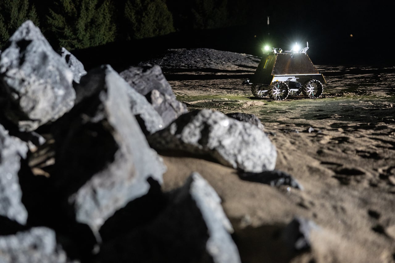 Rocks in the foreground, with a rectangular robot wheeling around on a sandy surface in the background.