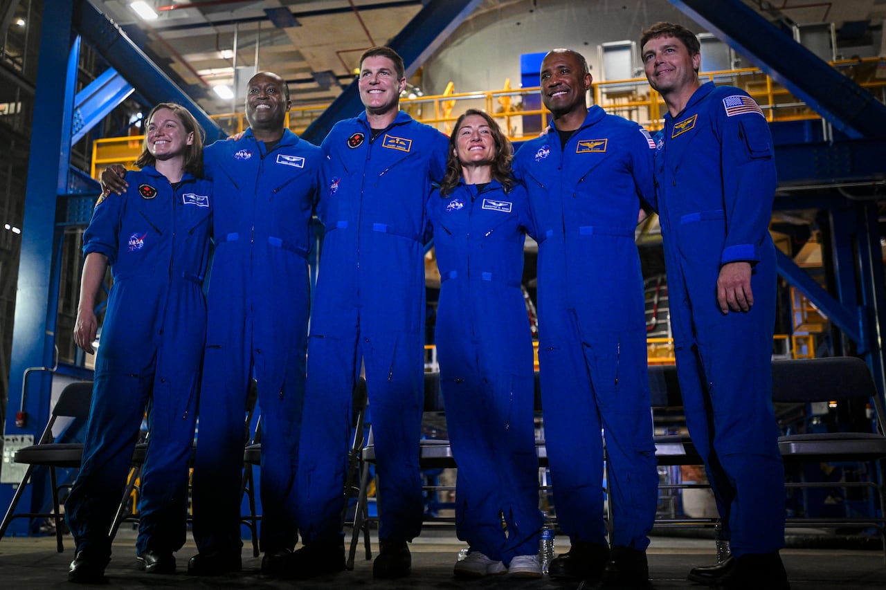 Six people stand with their arms around each other. They're wearing matching blue jumpsuits and are smiling for the camera.