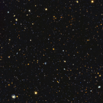 Image is filled with distant galaxies in colors of yellow-gold, red, blue, white. Lower left holds a face on spiral galaxy.