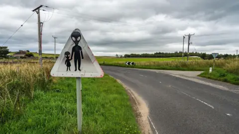 Alamy A road sign showing two aliens in silhouette crossing a road. They have large heads with huge almond-shaped eyes. The larger - presumably adult - alien is holding the hand of a small, presumably infant, alien. The sign is weathered, triangular, and by the side of a bending road with a small bungalow in the background.