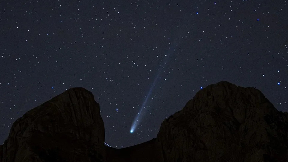  A white streak of a comet's tail can be seen in the center of the image as it streaks diagonally from top right to lower left between the dark silhouette of rocks blocking part of the starry night sky. 