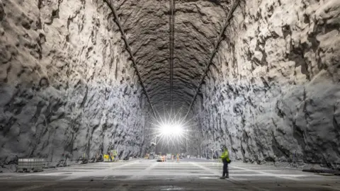 Matthew Kapust / SURF Workers in orange overalls in the background appear like tiny insects in a big white cave, illuminated with bright white electric lights that form two starbursts at the top of the frame. In the foreground a person wearing a green jacket, appearing larger in the frame, studies a laptop.