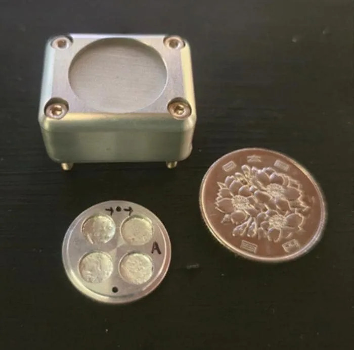 A small metal box with a circular cutout and two corresponding circular metal pieces next to it on a dark table