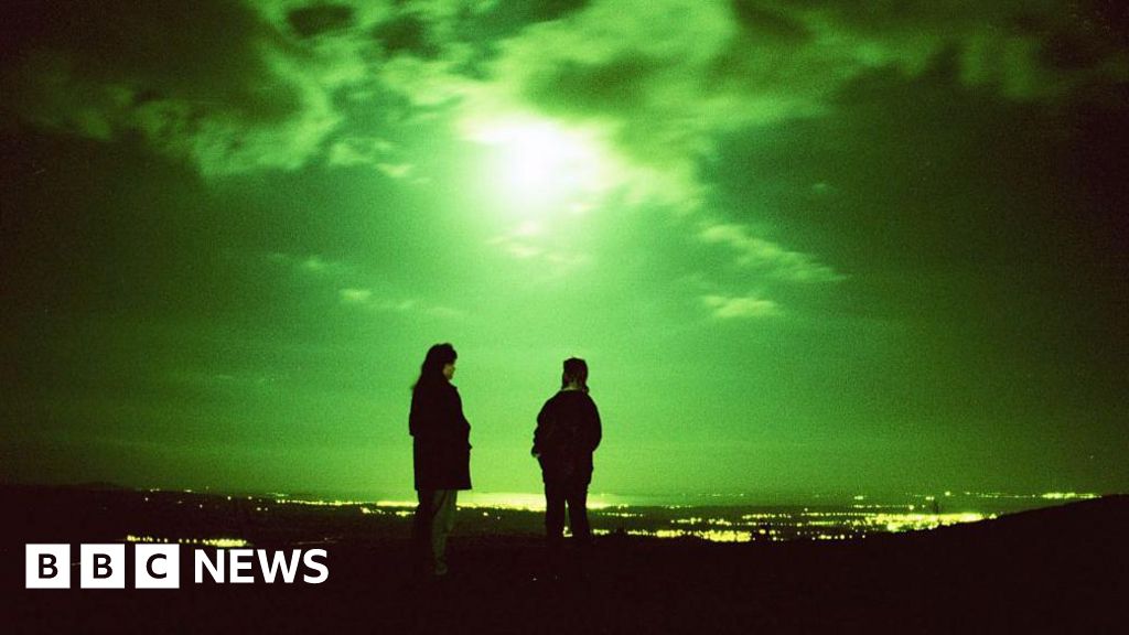 When Scotland was the world’s UFO hotspot Getty Images Two people standing with their backs to us at night. They are high up, overlooking a landscape of lights and water in the distance. The moon is high and the whole night sky is green-tinged and eerie.