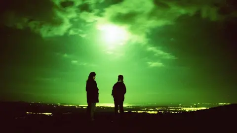 Getty Images Two people standing with their backs to us at night. They are high up, overlooking a landscape of lights and water in the distance. The moon is high and the whole night sky is green-tinged and eerie. 