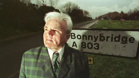 Getty Images Billy Buchanan, a man with white hair, is wearing a green tartan suit and standing in front of a weathered road sign which reads Bonnybridge A803. Houses can be seen in the background. He is looking up into the air.
