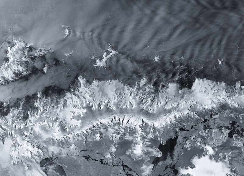 Satellite image showing a snow-covered mountain range bordered by dark, icy waters and streaked clouds. Jagged peaks and ridges dominate the landscape, contrasting sharply with the surrounding areas.