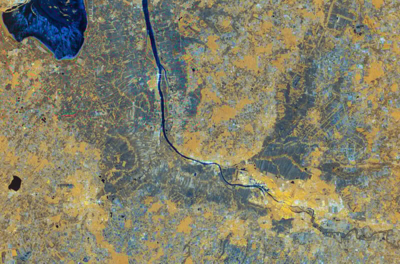 Satellite image of a river winding through a landscape with blue water features, tan and grey areas representing land, and visible patterns of fields, roads, and urban development.