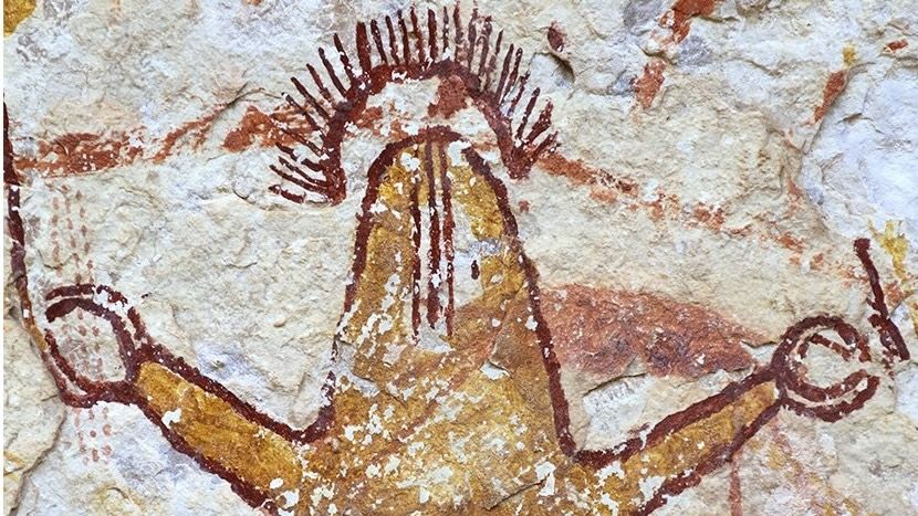 Red and yellow rock art of a figure with round fingers and outstretched hands.
