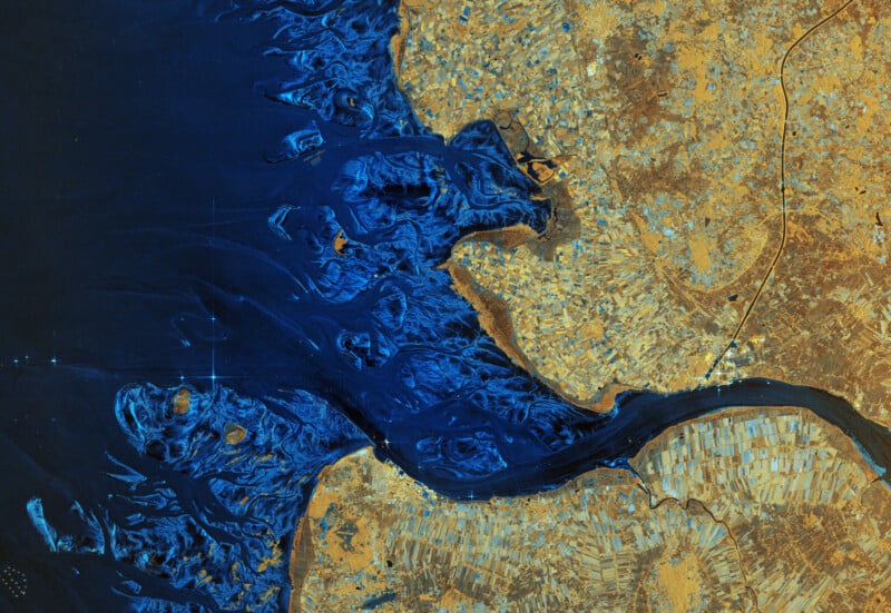 Satellite image shows a vivid blue river delta meeting golden-brown land, with branching waterways and agricultural fields visible along the coastline. Thin roadways curve through the landscape.