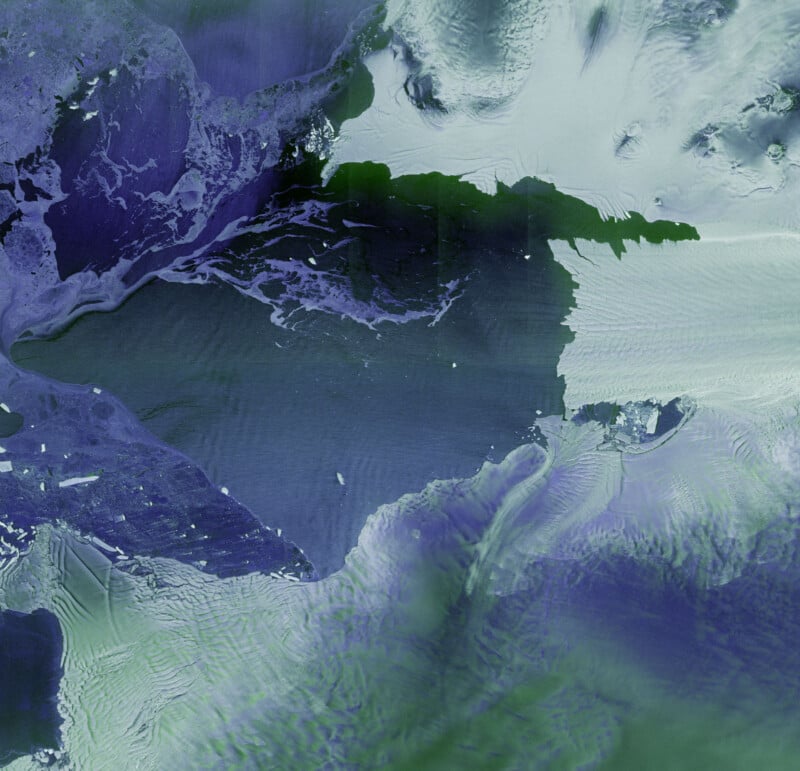 Satellite image showing a large, partially snow-covered landmass surrounded by icy water and floating ice. The landscape features shades of green, purple, and white, with textured patterns indicating ice and snow formations.
