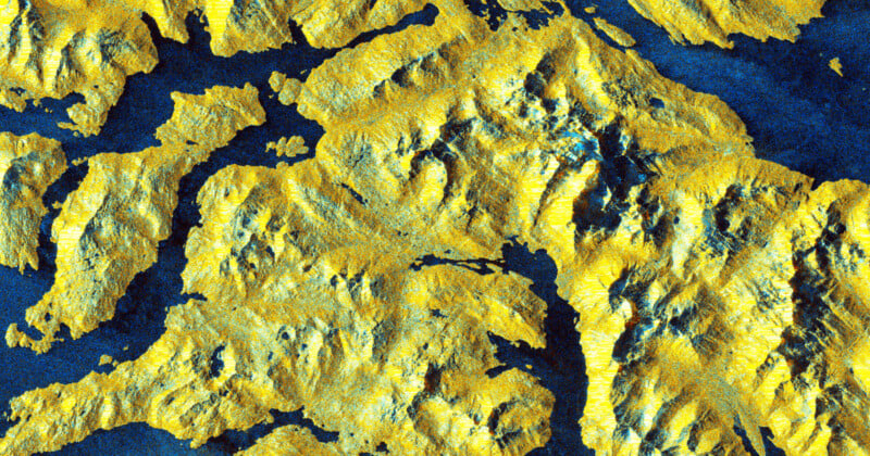 Satellite image showing a rugged, mountainous terrain with yellow and green elevations and deep blue valleys or waterways, highlighting the topography and varied landscape features.