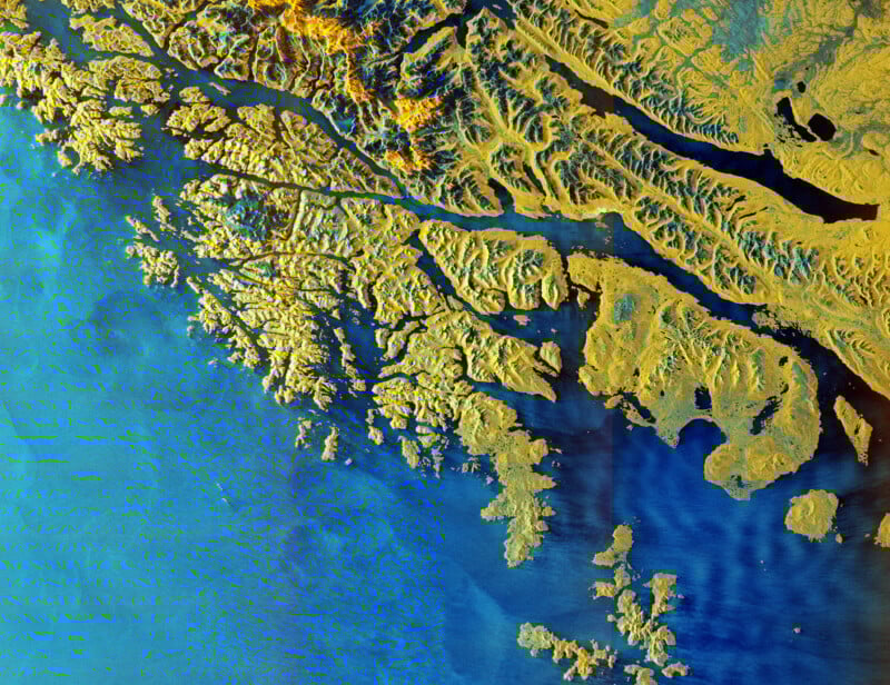 A vibrant satellite image shows a rugged coastline with yellow landmasses and blue water. Numerous peninsulas and inlets extend into the sea, highlighting intricate coastal geography.