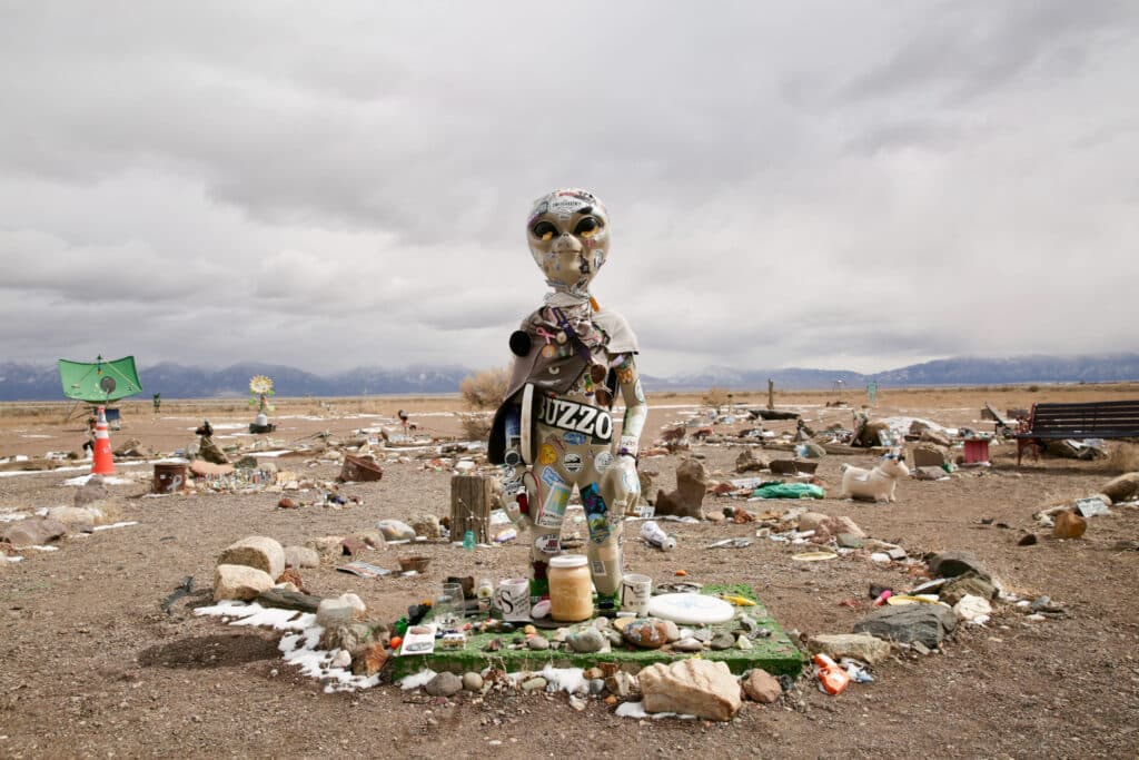 A tan alien statue stands in a rock garden under overcast skies.