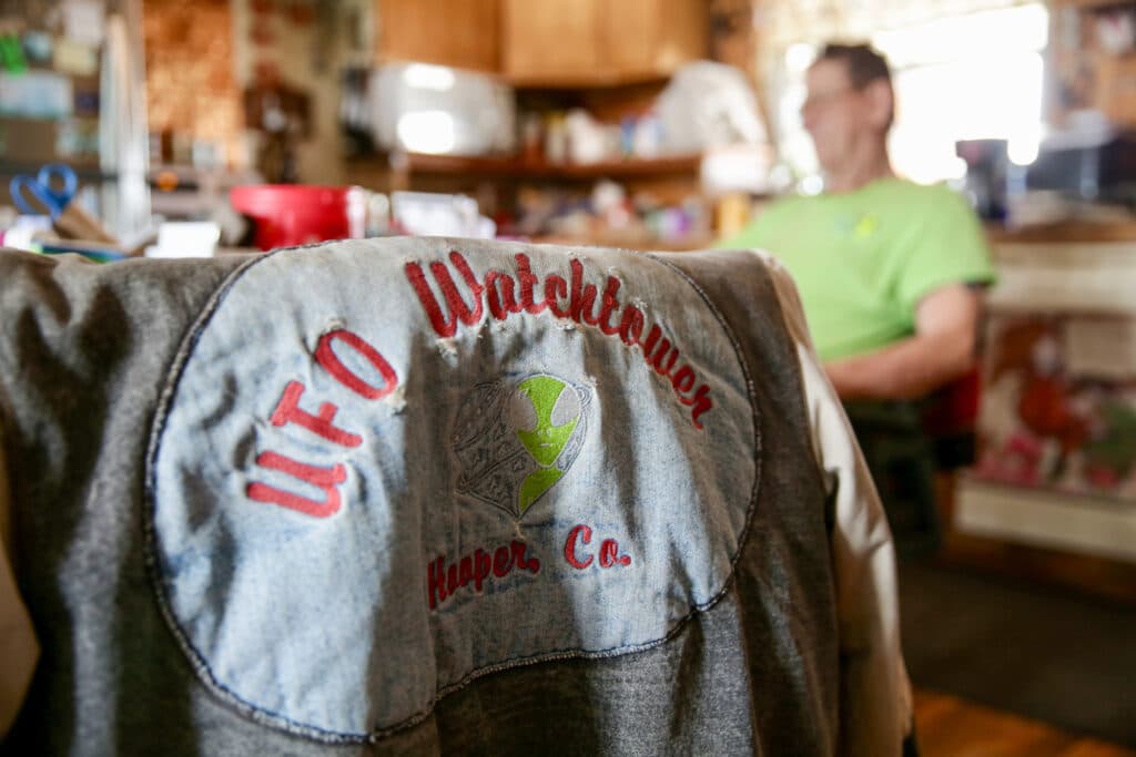 A denim jacket advertises the UFO Watchtower. Judy Messoline’s partner, Stan Becker, sits at the table in the background.