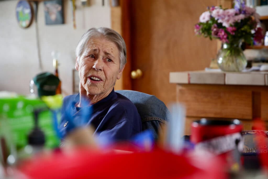 An elderly woman sits at a dining room table, speaking to someone not in the frame.