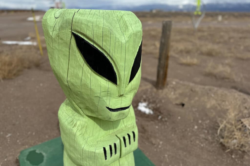 A green, carved wooden alien stands in the foreground of a gravel road.