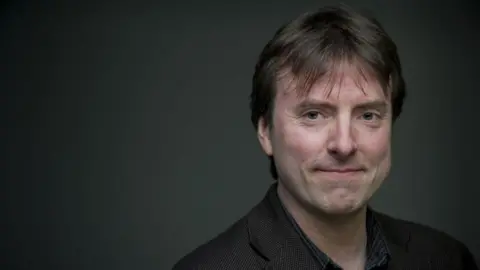 Sheffield Hallam University Dr David Clarke is a slim man in his 50s, with straight, dark hair. He is smiling. He is wearing a dark, dog-tooth check jacket and a dark striped shirt. He is standing against a dark grey, lit backdrop.