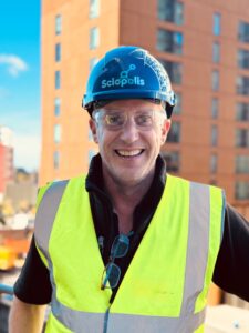 Charlie Mitchell with a hard hat on and a high visibility vest in front of a building smiling at the camera