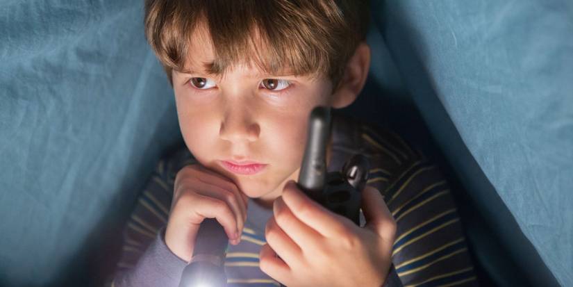 A young boy sits under his blanket with a flashlight and a walkie-talkie in DARK SKIES