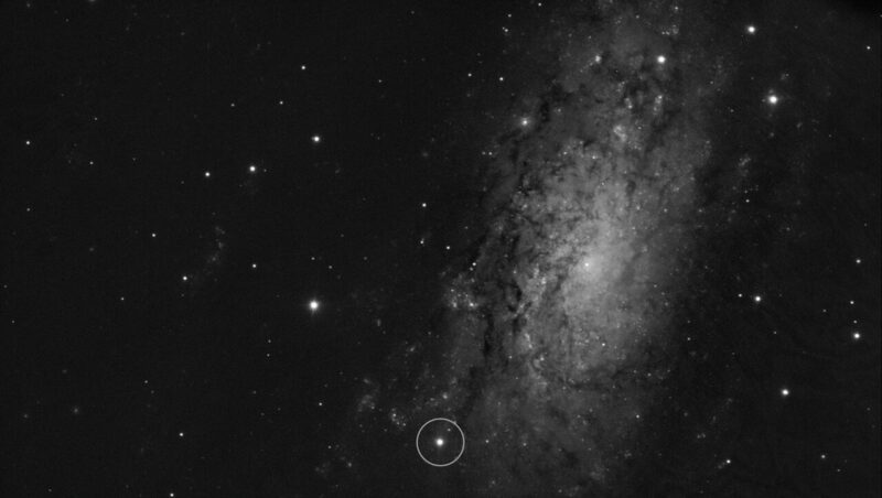 Black and white image of a spiral galaxy with a bright point of light near the bottom circled.
