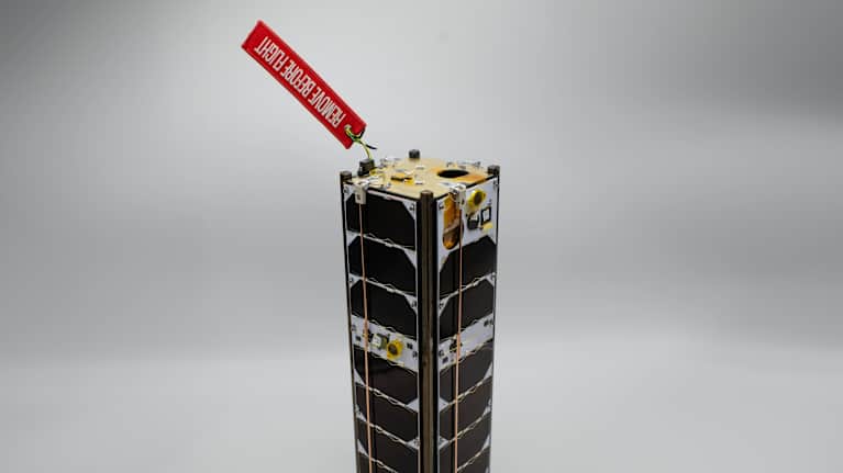 A rectangular shaped microsatellite on a white background, with a red tag attached to it that reads: "remove before flight".