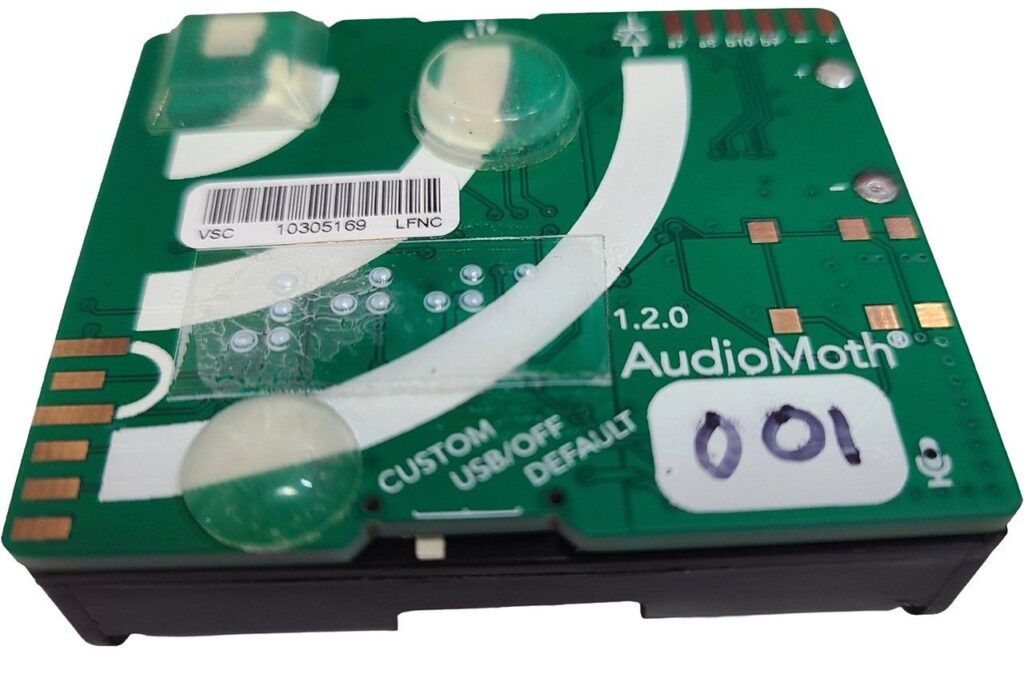 A small green circuit board, about half the size of a smartphone, attached to a pack of three AA batteries. The board has raised bump dots, printed circuit lines, small copper pads, a white barcode sticker, and a white label with the handwritten number “001” on it.