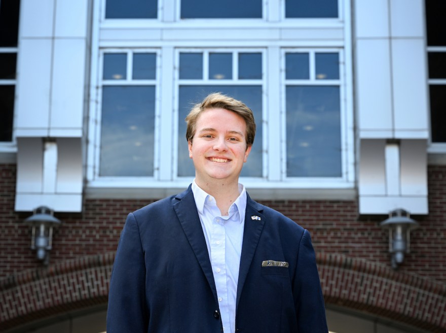 A student wearing a navy blazer and button-down shirt smiles while standing in front of a brick and white-paneled academic building with large windows.