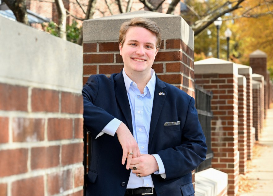 A student in a navy blazer leans against a brick column along a campus walkway lined with matching brick pillars and fall foliage.