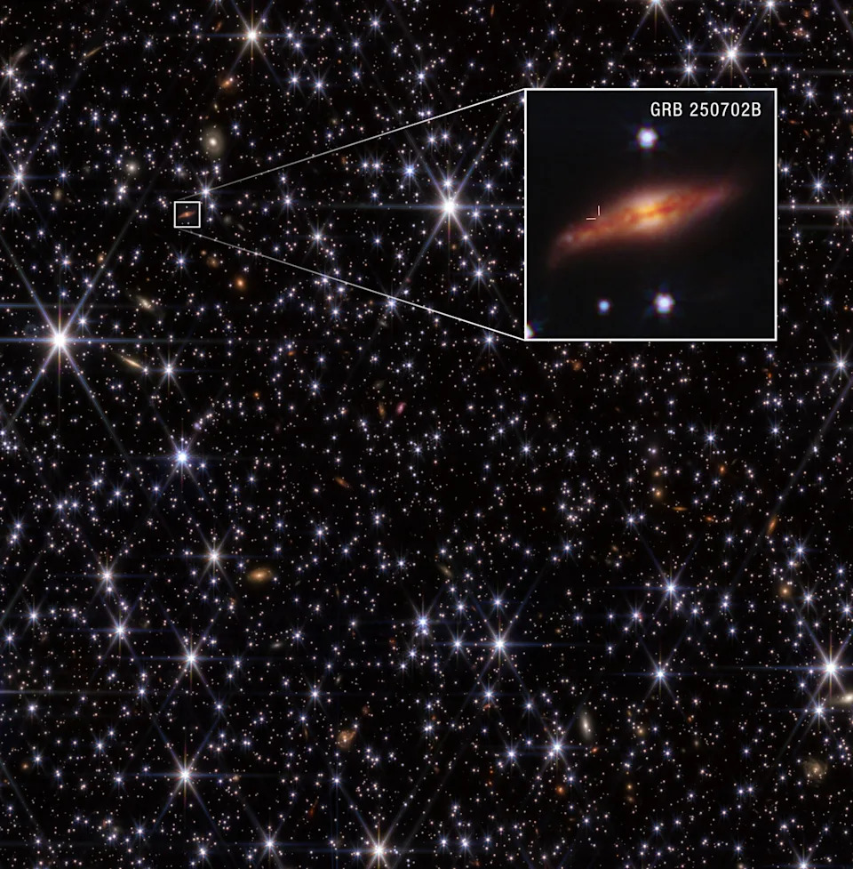 Against the blackness of space is a riot of white stars of varying brightness, with long diffraction spikes extending from the brightest ones. Also visible is a variety of cream-colored, orange, and brownish galaxies. A white box surrounds one galaxy in the upper left, located above a line joining the scene’s two brightest stars. Lines extend to the right from this box and lead to an enlarged inset showing the orange, edge-on disk galaxy in more detail. Horizontal and vertical lines pinpoint the burst’s location within its host galaxy. Text in the inset reads “GRB 250702B” in white