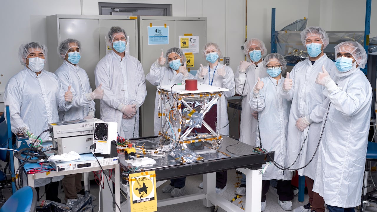 Nine people, dressed in PPE, stand in a clean room around a silver spacecraft giving thumbs ups.