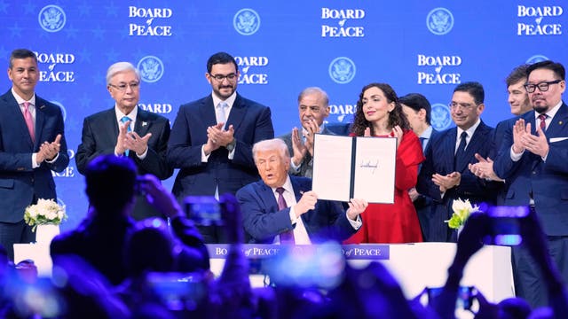 A seated Donald Trump holding up a charter during a signing ceremony while others stand behind him clapping