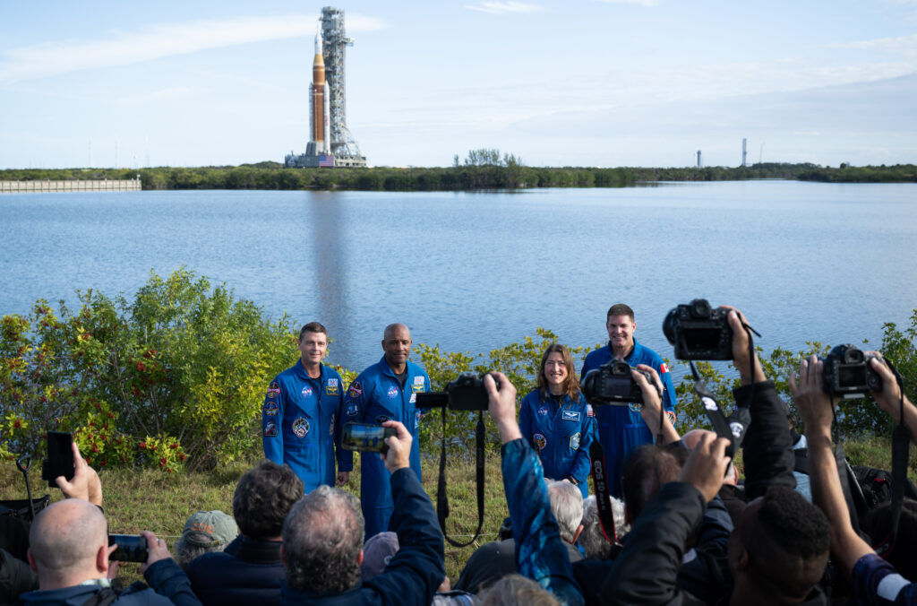 Astronauts take questions from members of the media with their rocket in the background. 