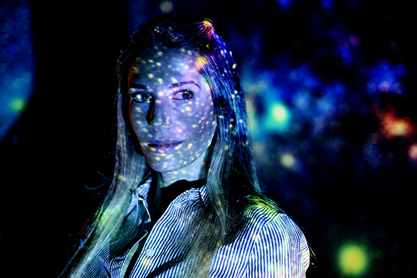 Portrait of Jacqueline McCleary in a dark room with a light setup that mimics the night sky projected onto her. 
