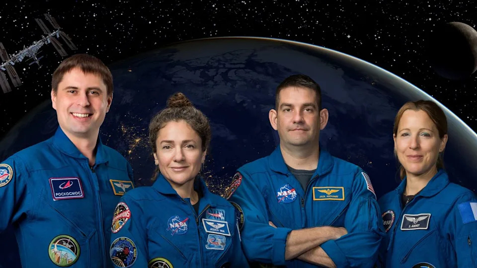  Four humans stand in blue jumpsuits in front of a background of a dark earth and shadowed moon, with a small ISS in the top left. . 