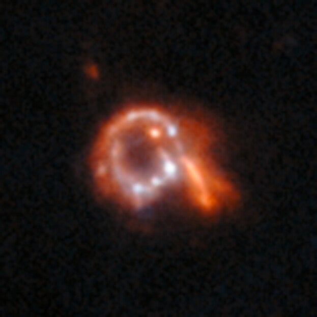 A small image of a mostly red galaxy. Unusually, it is ring-shaped with spots of light around the ring, a protruding arm on one side and a dark hole in the center.
