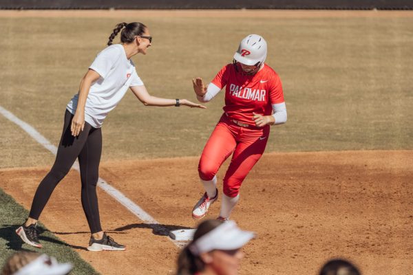 Palomar softball player Alexis Huey in a red uniform and white helmet high fives the outstretched arm of her coach while she rounds third base after hitting a home run to left field.