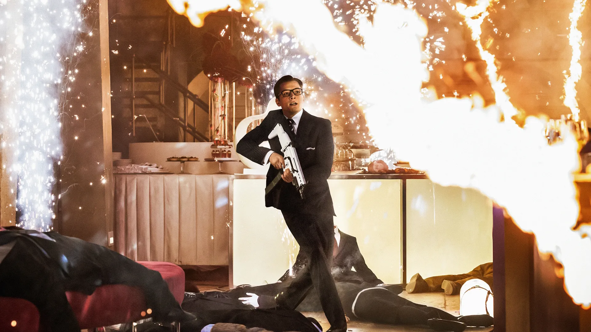 Eggsy (Taron Egerton), dressed in a suit and carrying a large gun, runs past an explosion