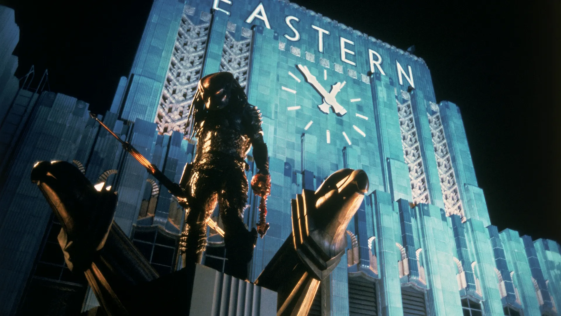 The Predator (Kevin Peter Hall) stands high up on a building at night, with the clock tower visible in the background