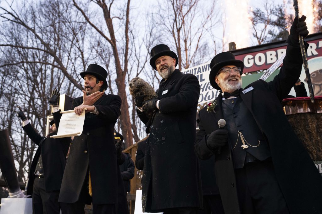 A series of men wearing all black with black top hats address a crowd off camera. The middle man holds a large brown rodent and the man on the left holds an open scroll