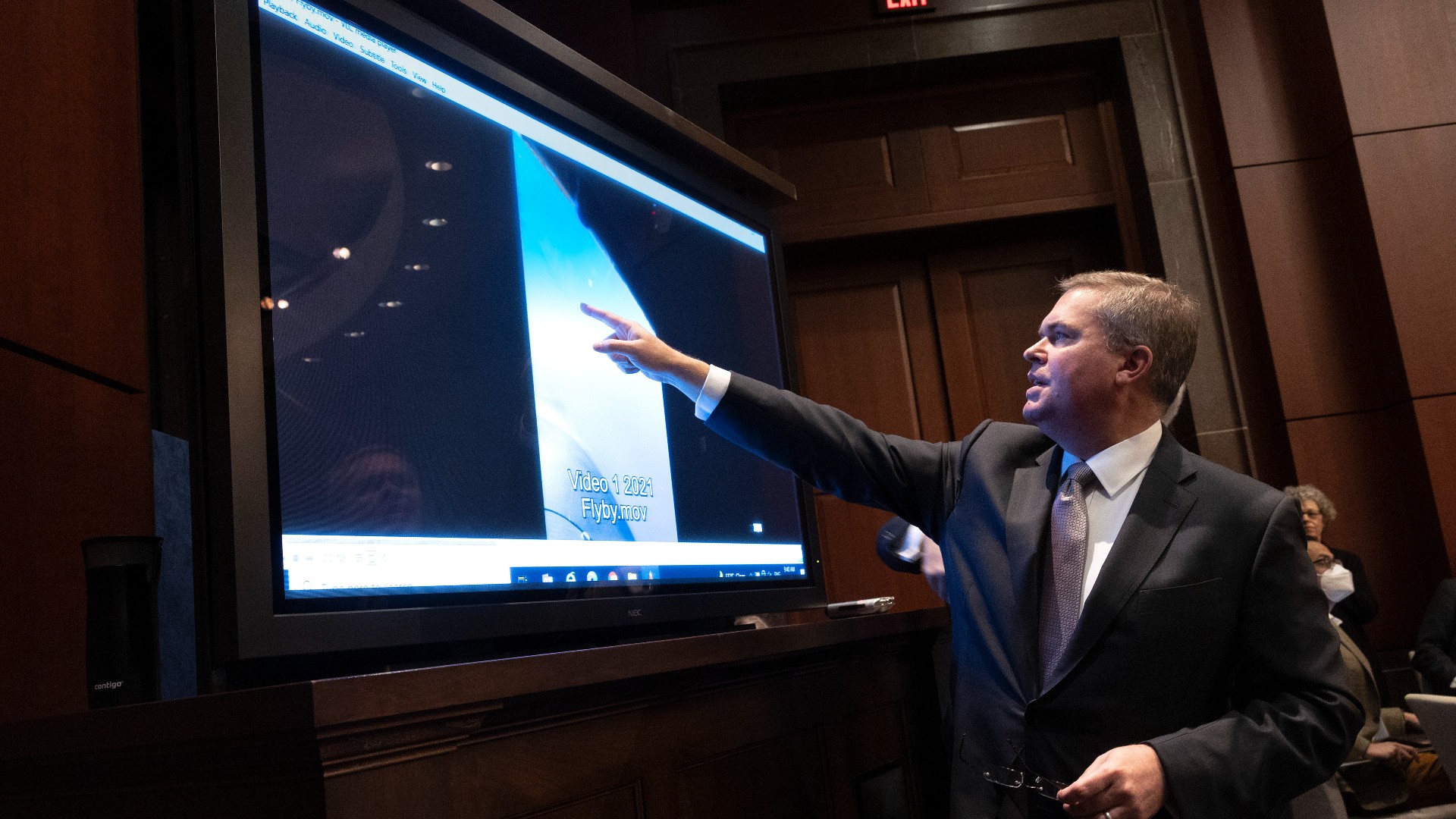 a man in a suit points at a video screen in a wood-paneled room