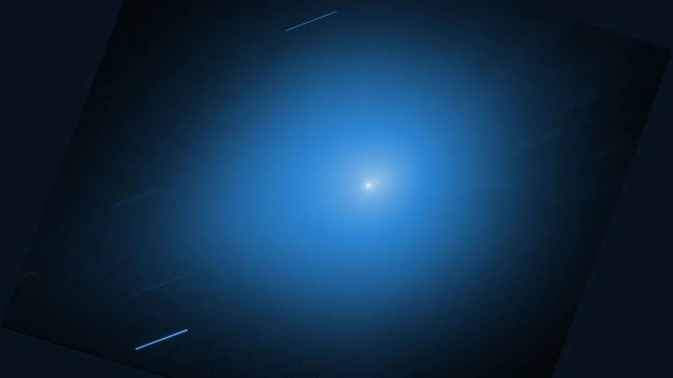  A white light of the comet 3I/ATLAS is surrounded by a blue glow against a black background. 