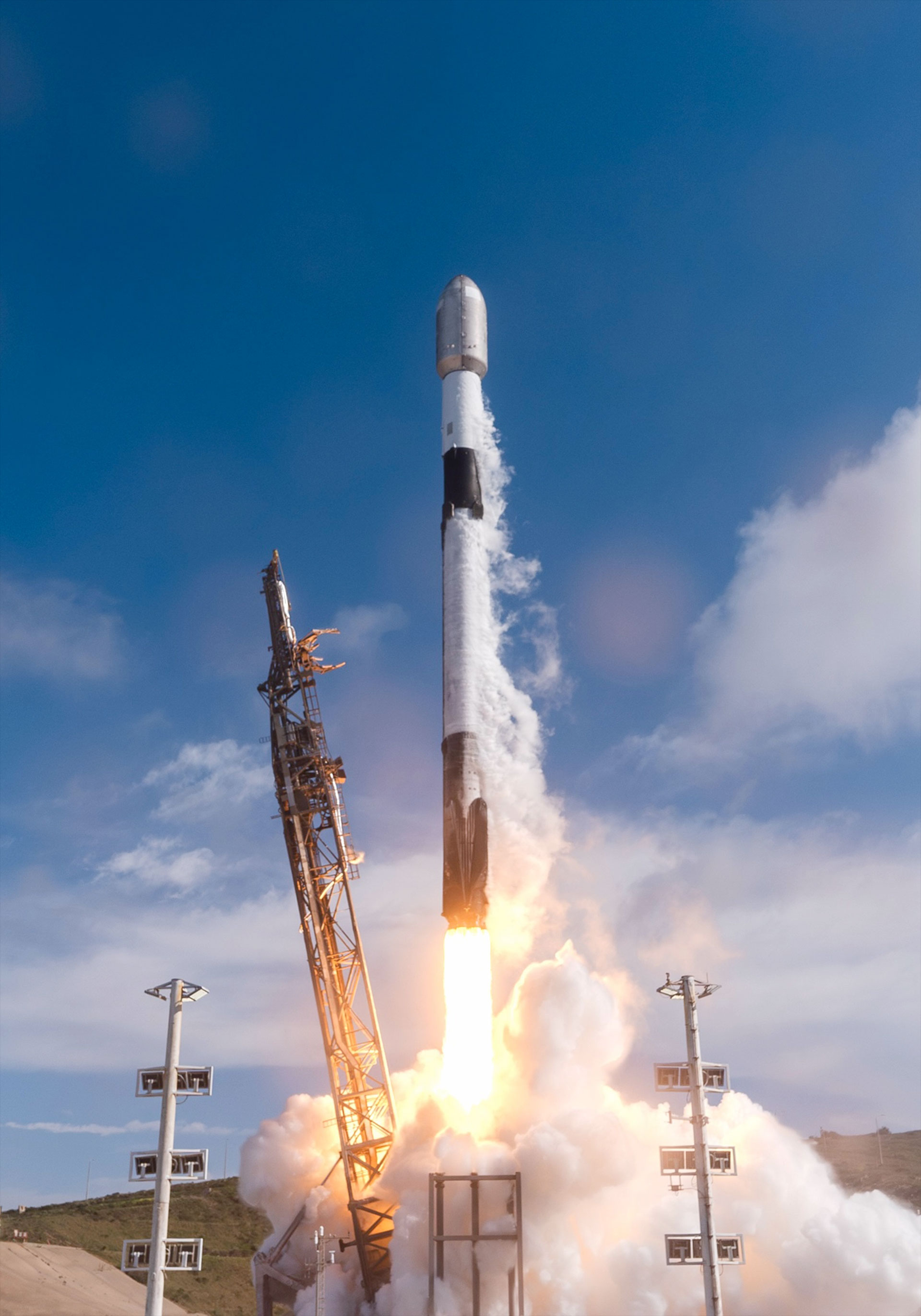 a white and black rocket lifts off into a partially cloudy sky. backdropped by an ocean.