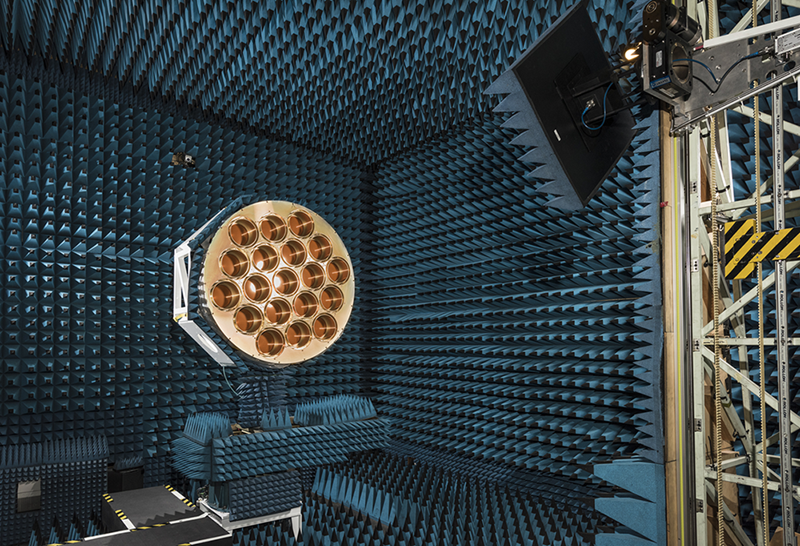 A large golden circle with 19 circular holes sits within a chamber covered in blue-grey foam wedges that absorb radio waves.