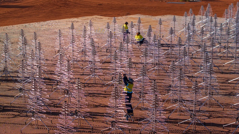 Three people in high-vis and sun-protective clothing installing a components on the top of silver-metal Christmas-tree shaped antennas, surrounded by even more antennas and the red dirt of Wajarri Yamaji Country in the Murchison.