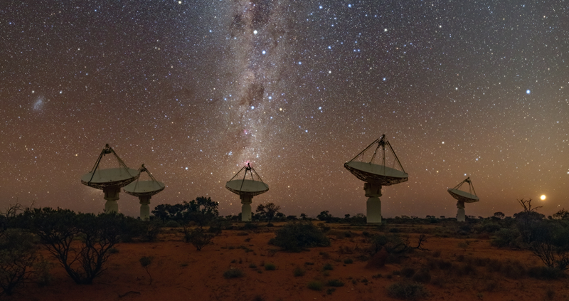 Five tall white dish-shaped radio telescopes point up to a star-filled night sky under the Milky Way. The Southern Cross and pointers are visible above the middle dish.