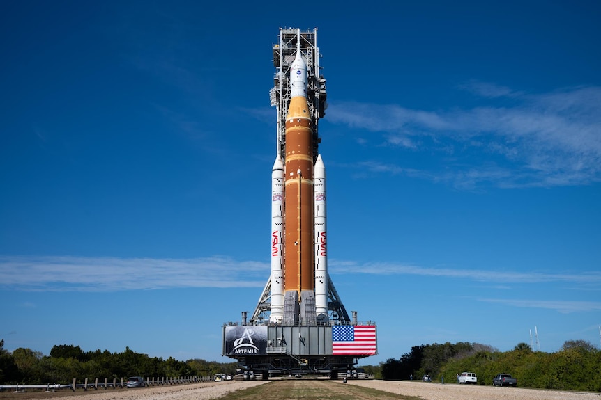 Large orange rocket flanked by two white fuel tanks and a white capsule on top on a trailer.