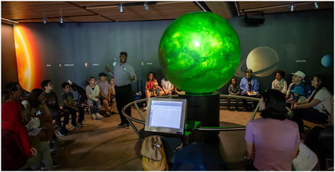 A group of people sit in a circular room, facing a large, glowing green sphere. A man stands talking in front of a backdrop displaying names of planets, such as Sun, Mercury, and Venus.