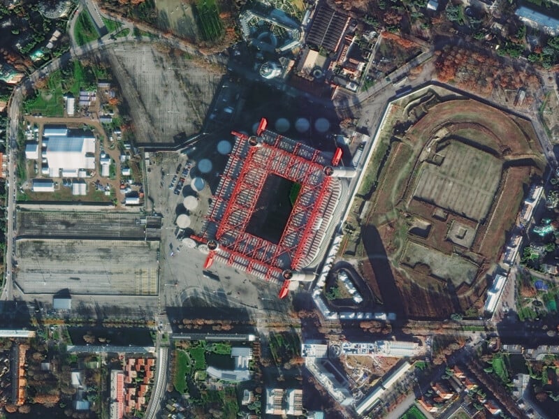 Aerial view of a large, rectangular stadium with a red roof and four corner towers next to an adjacent oval structure, surrounded by urban roads, buildings, and green spaces.