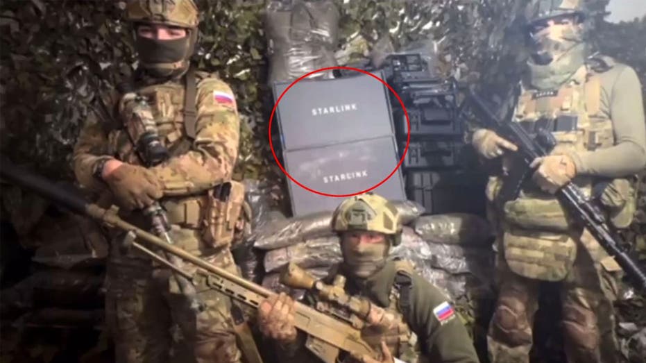 Three masked Russian soldiers in camouflage at a frontline position with two boxed Starlink units stacked behind them.